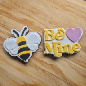 Valentines day cookies Be mine. 1 cookie is shaped as a bumble bee with black and yellow icing for the body with white iced wings. The other cookie is a pun, play-on-words showing, "Be Mine" coloured red on a white background. Next to it is a Pink love heard dividing the words
